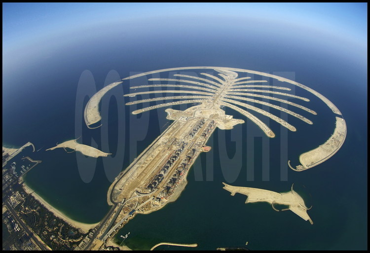Juin 2006. Chantier off shore du “Palm Jumeirah”. Avec ses 3 840 km2, Dubaï n’a qu’une courte façade littorale. Aussi, le petit émirat s’est-il lancé à partir de 2001 dans de pharaoniques projets touristiques, dans le dessein d’augmenter ses côtes en construisant les plus importantes îles artificielles du monde. Premier du genre, le « Palm Jumeirah » est un ensemble gagné sur la mer constitué d’un tronc et de 17 palmes cerclée par une digue longue de 11 km. Sur cet ensemble de 5 km de diamètre, 60 kilomètres de plages ont été créés en déplaçant plus de 100 millions de m3 de sable et de rochers. Inititialement prévu pour être achevé fin 2006, le chantier a pris du retard. Le « Palm Jumeirah» accueillera plusieurs dizaines d’hôtels, des milliers de maisons individuelles et d’appartements ainsi que des marinas, des restaurants, des parcs aquatiques, des centres commerciaux, des terrains de sport, des centres de thalassothérapie, des cinémas, etc. Un pont de 300 mètres reliera le tronc au continent, tandis qu'un monorail desservira le coeur du complexe, constitué d’un hôtel de 1 000 chambres et d’un parc aquatique.