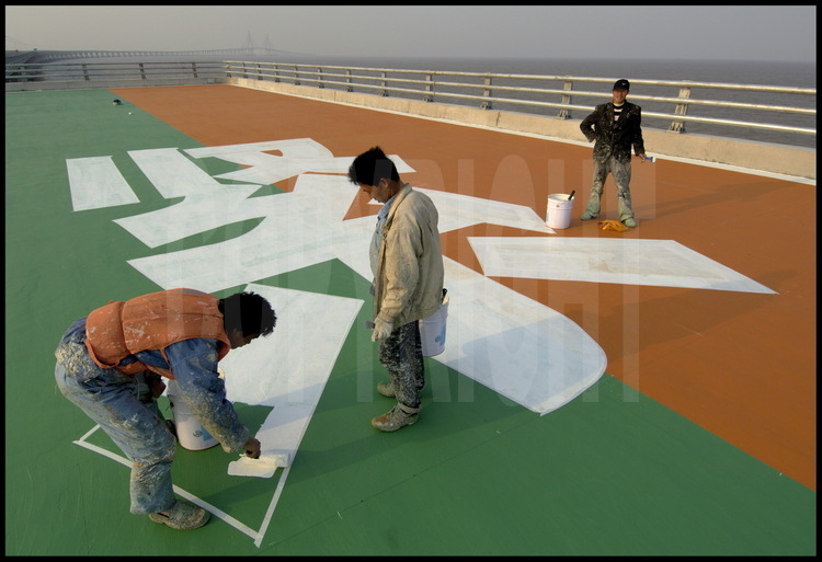 Last finishing works on Donghai bridge roadway at kilometric point 22. On a side platform, workers are painting ideograms meaning 
