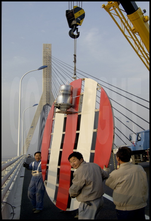 Last finishing works on Donghai bridge roadway at kilometric point 16 (main suspended bridge). Workers are installing, on the outside part of the roadway, signs for biggest vessels traffic. On background, northern tower of main suspended bridge (159 m high).