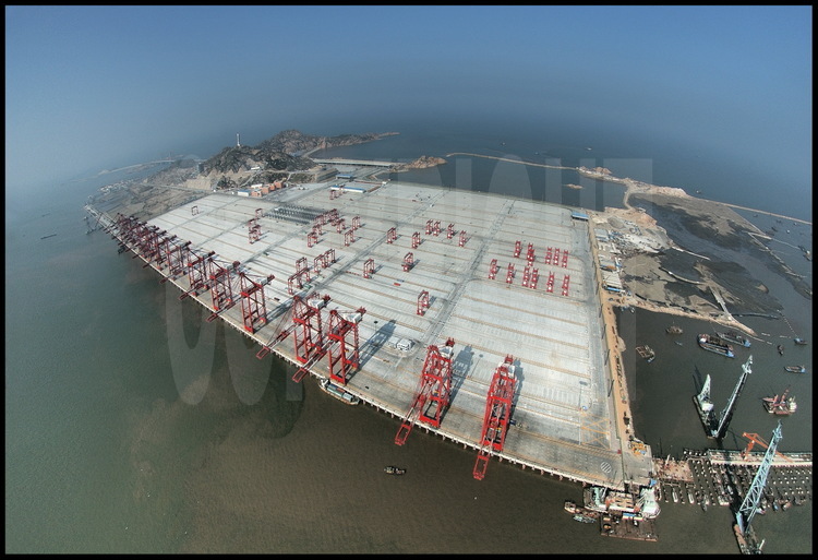 Along this 720000 meter square concrete slab, 1600 meter of wharf. Under, 2819 supporting piles. Above, the fifteen cranes of the first phase of the work, ready to meet world biggest vessels from december 2005. All working station are fully equiped with hi-tech radio recognition container localisation, which makes handling faster (less than 90 seconds for a full take off-landing one) and reduce service mistaking risk. These cranes are able to handle two containers in a row for a maxi weight of 65 tons and a 60 meter range. On left background, the end of Donghai bridge, Yangshan northern village and PC harbour center (glass building). On right foreground, installation of the supporting piles of the second phase of the work, ready by end 2006.