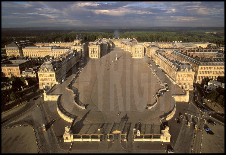 Vue aérienne du Château de Versailles. Au premier plan, l’entrée du château et la Grille Royale, puis la Cour des Ministres où trône la statue équestre de Louis XIV, la Cour Royale et la Cour de Marbre. En arrière plan, la grande perspective donnant sur le Grand Canal.