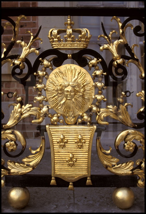 Sur la grille du balcon de la chambre du Roi, emblèmes royaux : tête d’Apollon, fleurs de lys, couronne fermée.