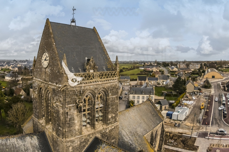 Normandie - Manche (50) - Sainte Mère Eglise : Site 6 : Sainte Mere Eglise. Le 5 juin 1944 a 23 heures, les forces alliees americaines sont parachutees au-dessus de Sainte-Mere-Eglise lors de l'operation Overlord. 15 000 hommes sont largues sur le village et ses environs, majoritairement issus de la 82e division aeroportee US (82nd Airborne), ainsi que de nombreux parachutistes de la 101e division aeroportee du fait d'erreurs de largage. Les Allemands tirent sur les parachutistes qui s'abattent sur le sol. John Steele, l'un d'eux, fut atteint au pied par une balle, ne pu controler son parachute et atterrit finalement sur le clocher de l'eglise vers 4h00 du matin. Il tenta de se liberer de son parachute tandis que sur la place, tout autour de l'eglise, la bataille faisait rage. Il prit alors la decision de faire le mort afin d'eviter de servir de cible a l'ennemi. Apres plus de deux heures, un soldat allemand vint le decrocher. Steele fut soigne et fait prisonnier, s'evada trois jours apres, rejoignit les lignes alliees et fut transfere vers un hopital en Angleterre. Depuis, un mannequin est accroche sur l'eglise de Sainte-Mere-Eglise en son hommage. John Steele est revenu plusieurs fois en Normandie pour les commemorations. Il est decede en 1969. Sainte-Mere-Eglise fut la premiere ville de France liberee par les airs. // France - Normandy - Manche (50) - Sainte Mère Eglise: Site 6: Sainte Mere Eglise. On June 5, 1944 at 11 p.m., the American allied forces parachuted above Sainte-Mere-Eglise during Operation Overlord. 15,000 men were dropped on the village and its surroundings, mainly from the US 82nd Airborne Division, as well as numerous paratroopers from the 101st Airborne Division due to drop errors. The Germans shoot at the paratroopers who fall to the ground. John Steele, one of them, was hit in the foot by a bullet, could not control his parachute and finally landed on the church tower around 4:00 a.m. He tried to free himself from his parachute while in the