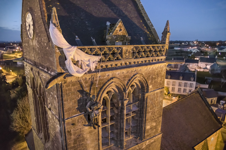 Normandie - Manche (50) - Sainte Mère Eglise : Site 6 : Sainte Mere Eglise. Le 5 juin 1944 a 23 heures, les forces alliees americaines sont parachutees au-dessus de Sainte-Mere-Eglise lors de l'operation Overlord. 15 000 hommes sont largues sur le village et ses environs, majoritairement issus de la 82e division aeroportee US (82nd Airborne), ainsi que de nombreux parachutistes de la 101e division aeroportee du fait d'erreurs de largage. Les Allemands tirent sur les parachutistes qui s'abattent sur le sol. John Steele, l'un d'eux, fut atteint au pied par une balle, ne pu controler son parachute et atterrit finalement sur le clocher de l'eglise vers 4h00 du matin. Il tenta de se liberer de son parachute tandis que sur la place, tout autour de l'eglise, la bataille faisait rage. Il prit alors la decision de faire le mort afin d'eviter de servir de cible a l'ennemi. Apres plus de deux heures, un soldat allemand vint le decrocher. Steele fut soigne et fait prisonnier, s'evada trois jours apres, rejoignit les lignes alliees et fut transfere vers un hopital en Angleterre. Depuis, un mannequin est accroche sur l'eglise de Sainte-Mere-Eglise en son hommage. John Steele est revenu plusieurs fois en Normandie pour les commemorations. Il est decede en 1969. Sainte-Mere-Eglise fut la premiere ville de France liberee par les airs. // France - Normandy - Manche (50) - Sainte Mère Eglise: Site 6: Sainte Mere Eglise. On June 5, 1944 at 11 p.m., the American allied forces parachuted above Sainte-Mere-Eglise during Operation Overlord. 15,000 men were dropped on the village and its surroundings, mainly from the US 82nd Airborne Division, as well as numerous paratroopers from the 101st Airborne Division due to drop errors. The Germans shoot at the paratroopers who fall to the ground. John Steele, one of them, was hit in the foot by a bullet, could not control his parachute and finally landed on the church tower around 4:00 a.m. He tried to free himself from his parachute while in the