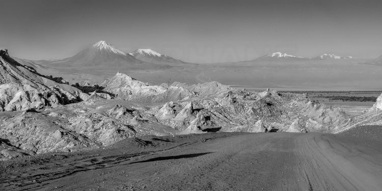 Chili - Désert d'Atacama : Valle de la Luna (altitude 2260 m.). Piste de sortie. En arrière plan, les volcans Licancabur (à g., altitude 5920 m.) et Juriques (au centre., altitude 5704 m.)