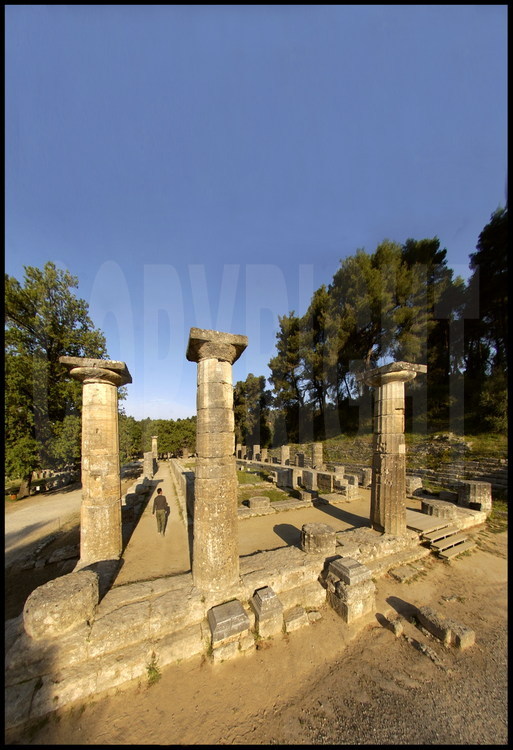 At the foot of Mount Kronion, the most sacred place of the Altis (the center of the sanctuary of Olympia) is occupied by the very ancient temple of Hera whose construction began in the 6th century B.C.  In the foreground, the restored columns seen from the North East.  In front of the Heraion, the Olympic spirit, born on this site, has spread today throughout the entire world.  Since the advent of the modern Olympic Games, the sacred flame, lit every four years, is carried from this temple to every corner of the world.