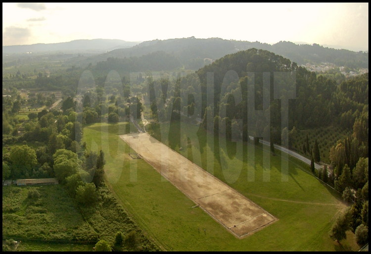 Aerial view of the stadium of Olympia.  In the background on the left, the sanctuary of Olympia.  On the right, the sacred hill of Mount Kronion.  The stadium’s current aspect dates back to the 4th century B.C.  The length of the track is 192.27 meters long, or 600 Olympic feet, which became the official distance of all ancient stadiums.  The spectators sat on the slopes on the ground, only the hellanodices’ (referees) stand on the east was made of stone.  45 000 spectators could sit on the stadium’s slopes to watch the competitions.