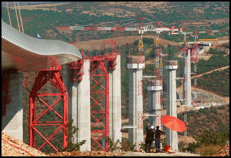 From the south bank (Larzac Limestone Plateau), view of the launching of the 4200 tons of metallic roadway and the 7 concrete piers in the final phase.  Using a GPS measurement system, recorded quantities come within a centimeter of the exact value.  The bridge’s astonishing curvature is due to the roadway’s considerable slant in the last meters of the operation.  In red, the rows of piers, temporary supports on which the roadway rests during the launching phase.  In the background, the Red Limestone Plateau.