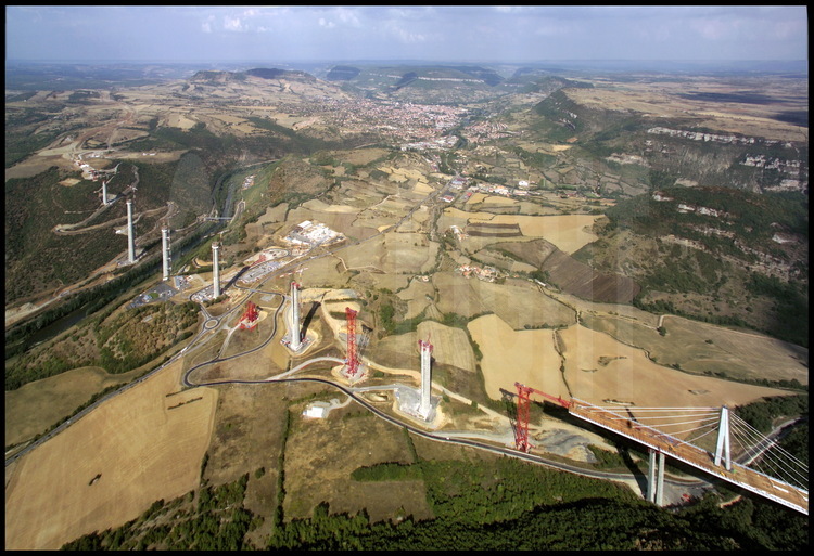General view of the viaduct’s construction site in June, 2003.  The first of seven parts of the metallic roadway in the launching stage from the southern bank of the Tarn valley.  Throughout this critical stage, in which calculations must be extremely precise, the roadway is “pushed” in the center of the valley before a new part of the roadway comes to push it to the next pier, 342 meters ahead.  In the background on the left, the Red Limestone Plateau.  In the center, the city of Millau.  On the right, the Larzac Limestone Plateau.