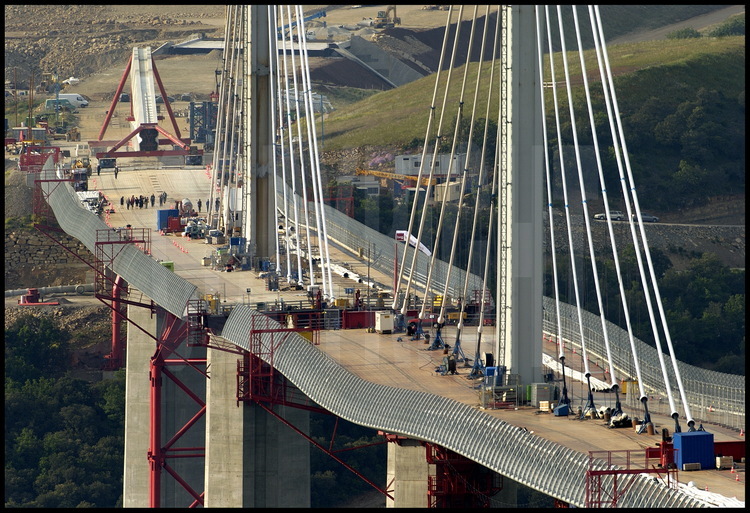 On top of the piers, 90-meter-high pylons will hold up the stays which will give the bridge its rigidity.  In the meantime, the roadway’s flexible steel seems to undulate between its different support points (171 meters apart) which are the piers and the temporary intermediary rows of piers.  In the background, the pylon which will top pier P1 is already under construction.