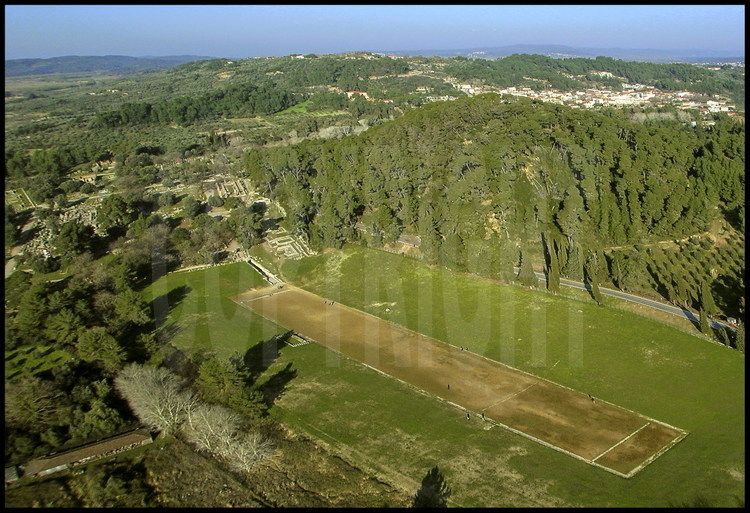 Aerial view of the ancient stadium of Olympia.  Its length of 192 m, useful for races, will become a model for all ancient stadiums (192m=1 stadium).  On the left, the sanctuary of Olympia, on the right, Mount Kronion which was sacred during Antiquity.  In the background on the right, the modern city of Olympia.